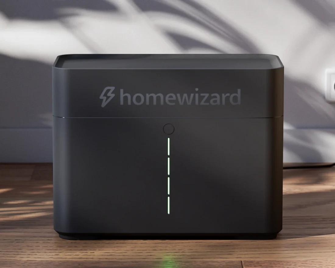 Een zwarte stekkerbatterij van HomeWizard op een houten vloer, met schaduw van planten op de muur erachter.