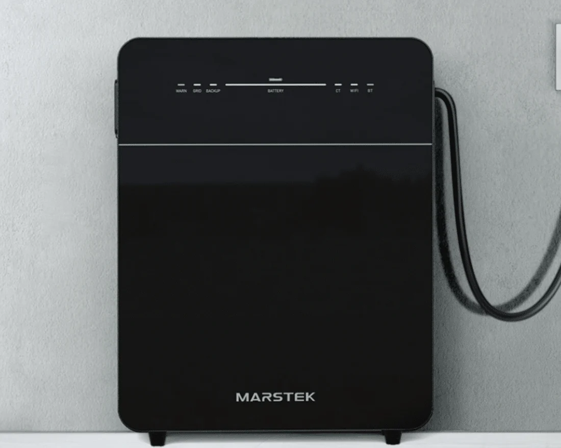 Zwarte Marstek Venus E 3.0 tegen een grijze betonmuur, met een dikke kabel richting stopcontact.